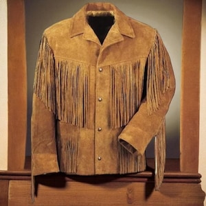 Op de afbeelding: Een bruin suède jasje met franjes op de borst, rug en mouwen. De jas heeft een knoopsluiting en een kraag. De jas wordt gepresenteerd op een houten oppervlak.