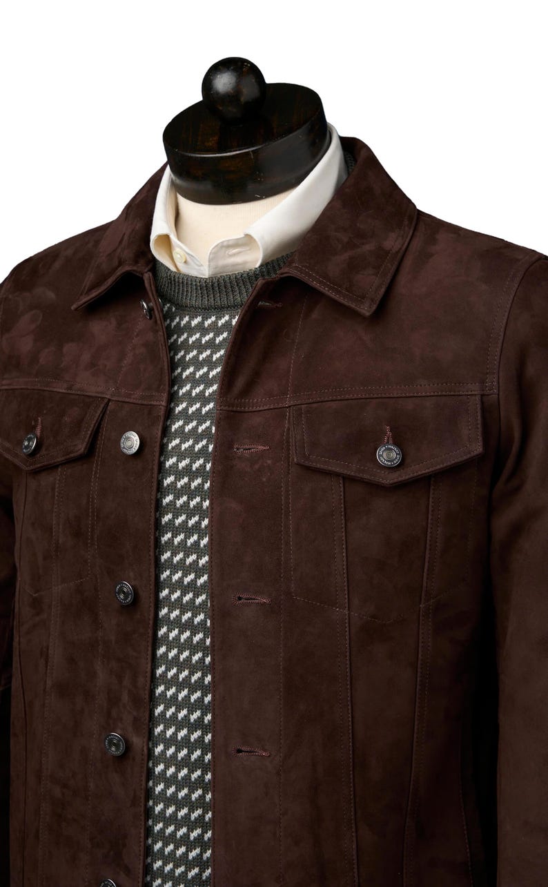 Op de afbeelding: Een bruin su&egrave;de jack met een knoopsluiting en twee klepzakken. Het jack wordt gedragen over een wit shirt en een grijze trui met patroon.