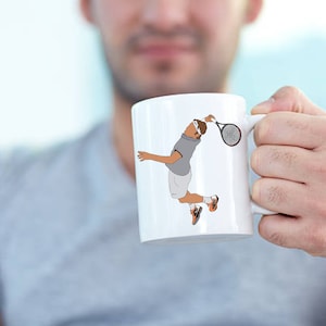 May include: White ceramic mug featuring a tennis player in action, with a gray shirt, white shorts, and a brown headband. The player is mid-swing with a tennis racket. The mug is held by a person.