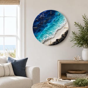 May include: A circular wall clock featuring a high-resolution print of an ocean shoreline with deep blue and turquoise water, white sea foam, and a sandy beach. The clock face includes thin, gold-colored hour markers positioned around the perimeter.