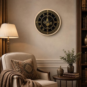 Könnte beinhalten: Runde Wanduhr mit schwarzem Zifferblatt und goldenen römischen Ziffern. Die Uhr hat ein verziertes Golddesign und arabische Kalligraphie in der Mitte. Sie ist an einer beigen Wand in einem Raum mit Lampe, Stuhl und Beistelltisch angebracht.