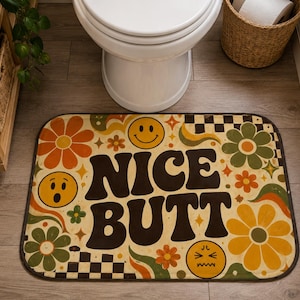 May include: A rectangular bathroom rug featuring a cream background with a retro-inspired pattern of orange, yellow, and green flowers, checkered squares, and three yellow smiley faces. The text NICE BUTT is printed in large, bold, dark brown letters.