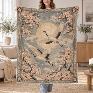 May include: A rectangular throw blanket featuring a printed design of two white cranes in flight against a large pale yellow moon. The scene is framed by blooming pink cherry blossom branches on a muted blue background with a decorative border.