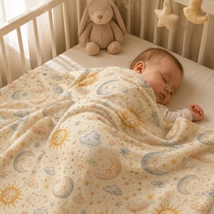 May include: A cream-colored baby blanket featuring a repeating pattern of smiling suns, crescent moons, stars, and soft blue and pink clouds. The blanket is draped over a sleeping infant in a crib, with a plush bunny toy visible in the background.