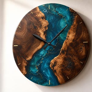 May include: A round wall clock featuring a natural wood grain face with a central, flowing river-like inlay of translucent teal and blue resin. The clock has thin, gold-colored hour markers and two black hands positioned at the center of the dial.