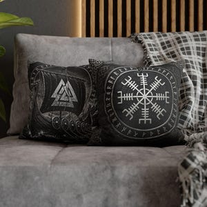 Op de afbeelding: Twee zwart-witte decoratieve kussens met Viking-ontwerpen. Eén kussen toont een Vikingschip en een driehoekig symbool, de andere een cirkelvormig embleem met runen. Ze liggen op een grijze bank en geven een vleugje Noorse mythologie.
