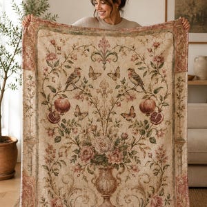 May include: A rectangular throw blanket featuring a cream-colored background with a central urn filled with pink flowers. The design includes symmetrical branches with birds, butterflies, and pomegranates, framed by a decorative floral border.