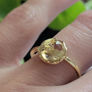Puede incluir: Un anillo de oro con una gema amarilla redonda. El anillo tiene una banda delgada y un engaste de bisel. La gema es translúcida con inclusiones internas. El anillo se lleva en un dedo.