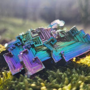 May include: A close-up of a colorful bismuth crystal formation. The crystal displays a vibrant array of colors, including blues, greens, purples, and yellows. The geometric structure of the crystal is clearly visible, with a mossy background.