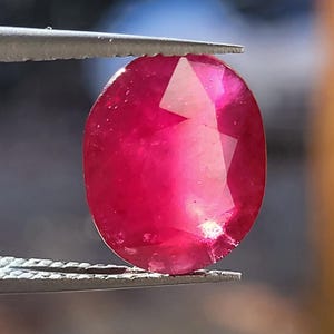May include: A faceted, oval-shaped ruby gemstone held by tweezers. The ruby displays a deep red color with internal light reflections. The gemstone is a focal point, showcasing its clarity and cut.