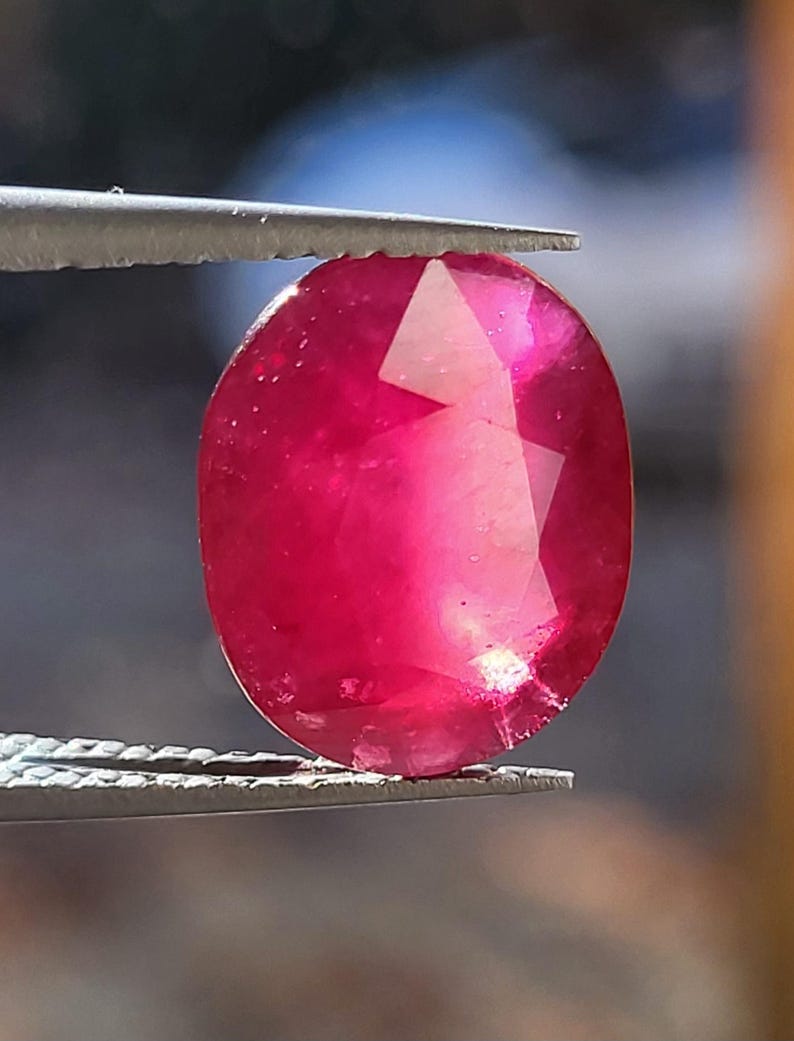 Op de afbeelding: Een gefacetteerde, ovaalvormige robijn edelsteen, vastgehouden door een pincet. De robijn toont een dieprode kleur met interne lichtreflecties. De edelsteen is een blikvanger en toont zijn helderheid en slijpsel.