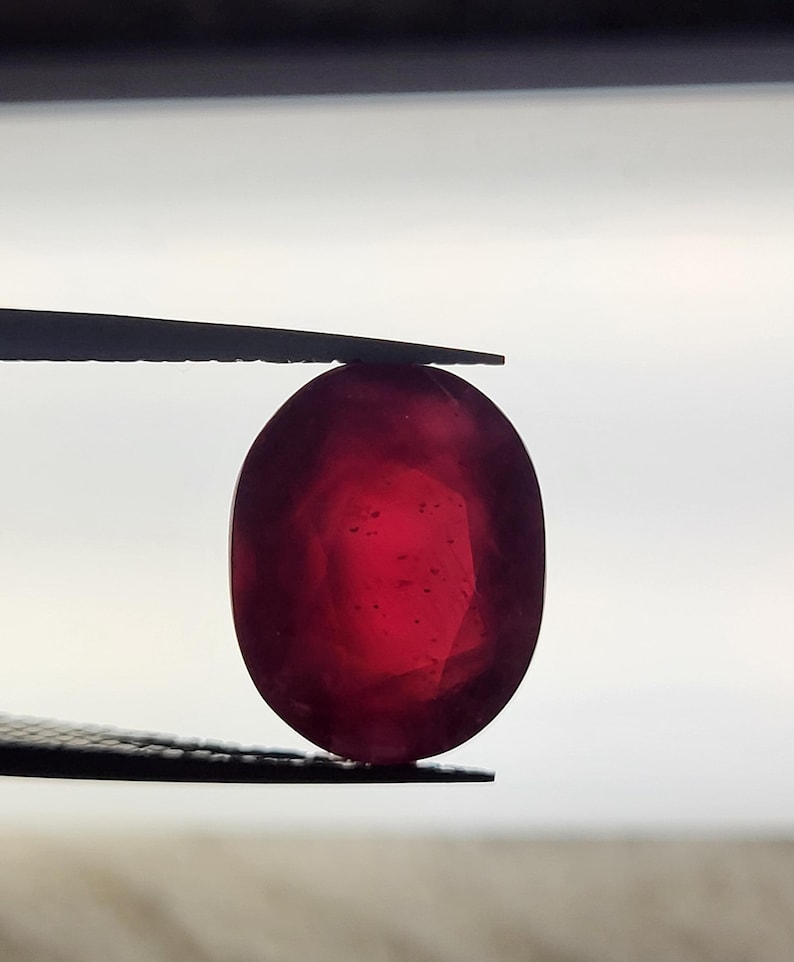 Op de afbeelding: Een levendige, ovaal geslepen robijn, vastgehouden door een pincet tegen een heldere achtergrond. De dieprode steen vertoont interne facetten en een gepolijst oppervlak, wat de natuurlijke schoonheid benadrukt. Een close-up benadrukt de helderheid en kleur.