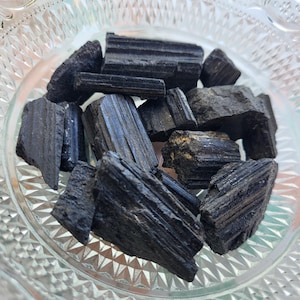 May include: A collection of rough, black tourmaline crystals in a clear, decorative glass bowl. The crystals have a striated texture and varying shapes, reflecting light. The bowl has a detailed, geometric pattern.