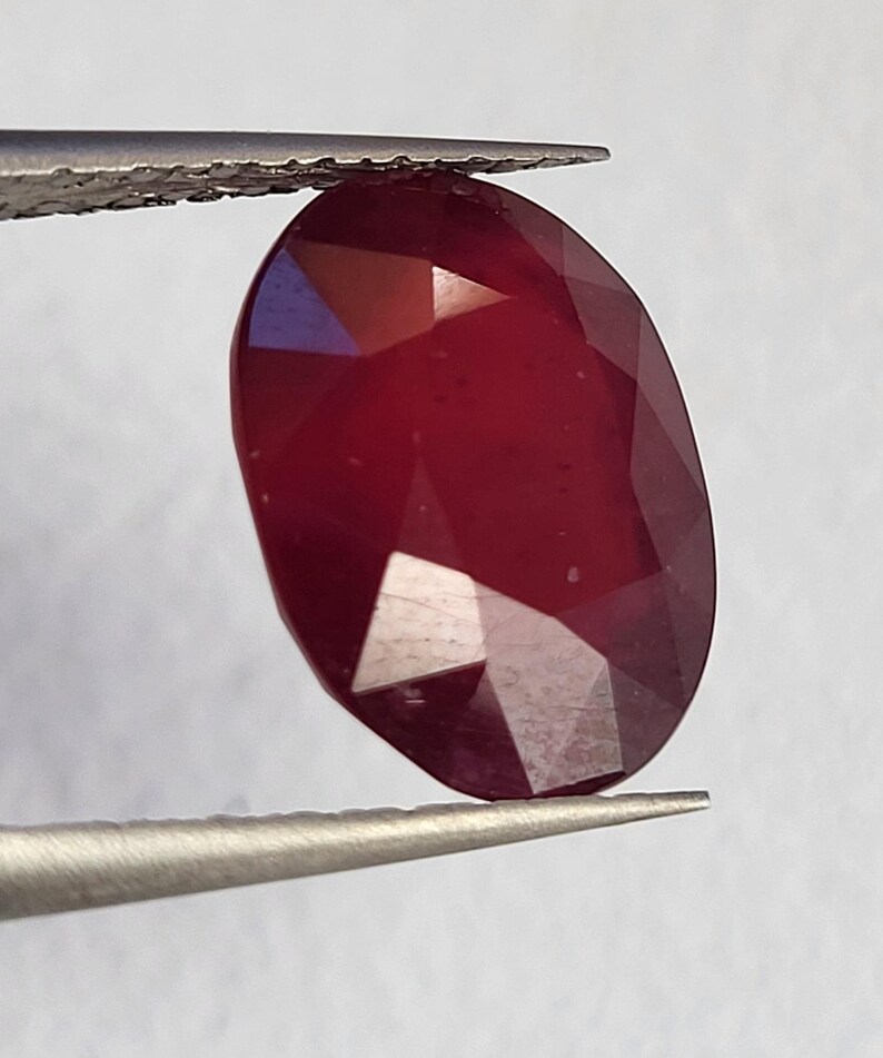 Op de afbeelding: Een gefacetteerde, ovaalvormige robijn edelsteen, vastgehouden door een pincet. De robijn heeft een diepe, rijke rode kleur met meerdere facetten die het licht vangen. De edelsteen wordt gepresenteerd tegen een neutrale achtergrond.