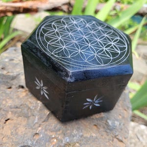May include: A black wooden box with a carved flower of life design on the top. The box has a hexagonal shape and is decorated with small white flowers on the sides.