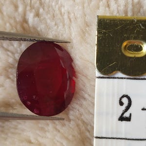 Op de afbeelding: Een gefacetteerde, ovaalvormige, dieprode edelsteen wordt vastgehouden door een pincet. Een meetlint is naast de edelsteen geplaatst en toont metingen van 5,08 cm en 7,62 cm. De edelsteen ligt op een zachte, getextureerde achtergrond.