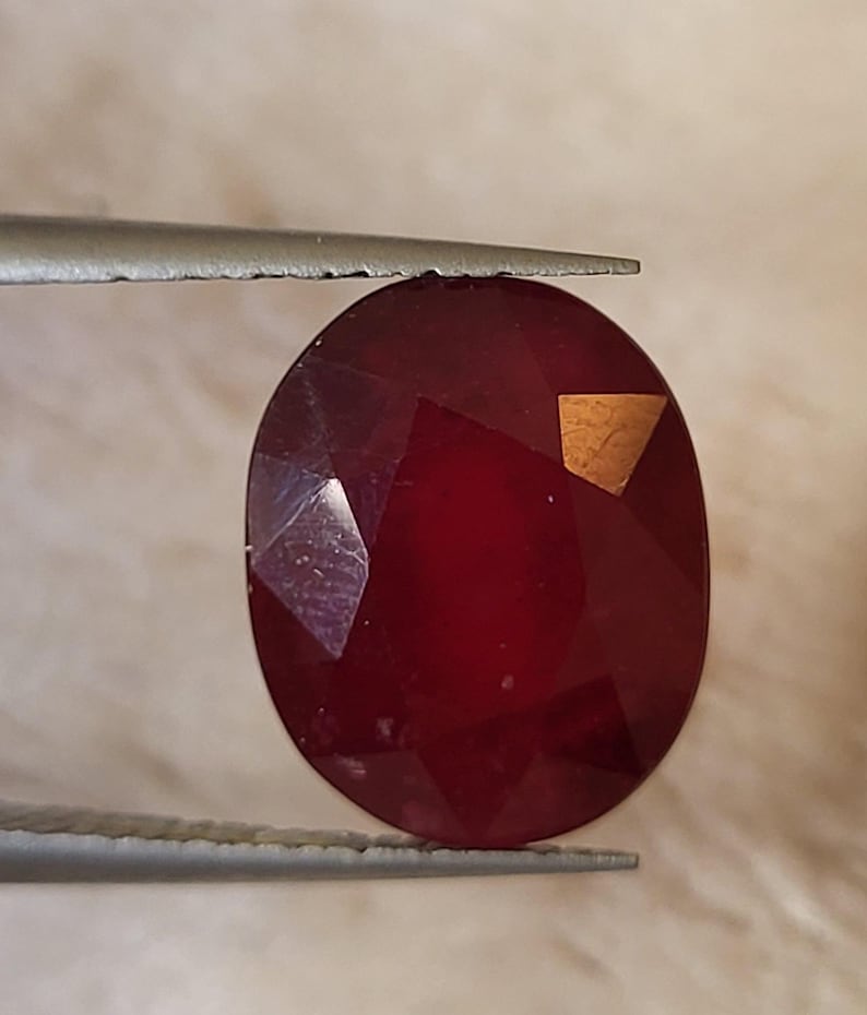 Op de afbeelding: Een gefacetteerde, ovaalvormige, dieprode edelsteen wordt vastgehouden door een pincet. De edelsteen heeft een gepolijst oppervlak met meerdere facetten die licht reflecteren. Een kleine, goudkleurige vlek is zichtbaar.
