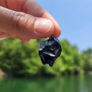 Peut inclure: Un petit pendentif en obsidienne noire de forme irrégulière. Le pendentif est suspendu devant un ciel bleu et un feuillage vert. L'obsidienne a une surface brillante et réfléchissante.