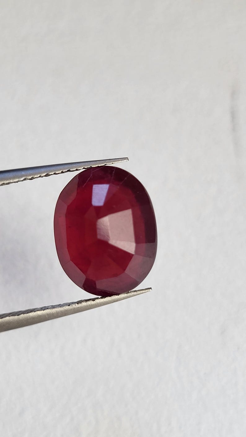Op de afbeelding: Een gefacetteerde, ovaalvormige robijn edelsteen, vastgehouden door een pincet. De robijn heeft een diepe, rijke rode kleur, met meerdere facetten die het licht vangen. De pincet is zilverkleurig en houdt de edelsteen tegen een witte achtergrond.