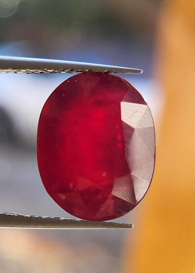 Op de afbeelding: Een gefacetteerde, ovaalvormige robijn edelsteen, vastgehouden door een pincet. De edelsteen heeft een diepe, verzadigde rode kleur, met facetten die het licht vangen. De achtergrond is een vage mix van oranje en blauw.