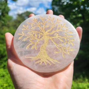 May include: A round, light pink stone with a gold-colored tree of life design. The stone is held in a hand against a backdrop of green foliage and a blue sky with clouds. The tree design is etched into the stone.