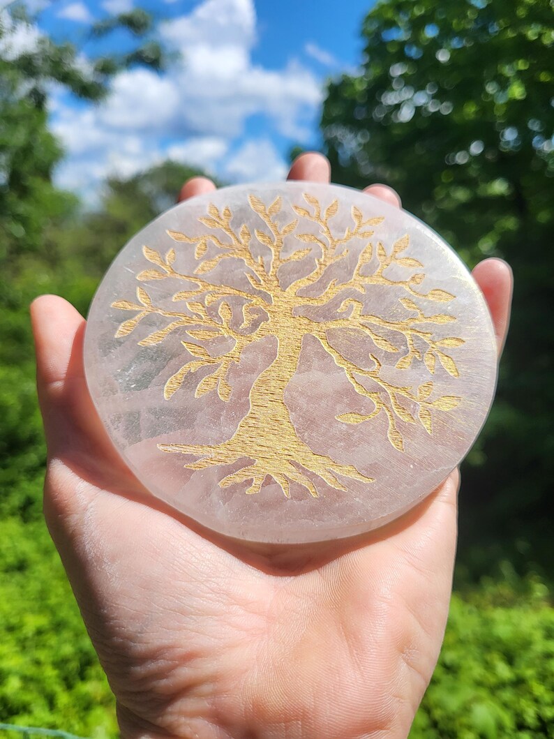 May include: A round, light pink stone with a gold-colored tree of life design. The stone is held in a hand against a backdrop of green foliage and a blue sky with clouds. The tree design is etched into the stone.