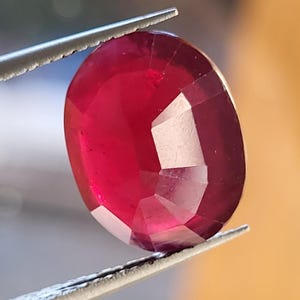 Op de afbeelding: Een gefacetteerde, ovaalvormige robijn, vastgehouden door een zilveren pincet. De dieprode edelsteen heeft meerdere facetten die het licht vangen. De afbeelding benadrukt de rijke kleur en het gepolijste oppervlak van de robijn.