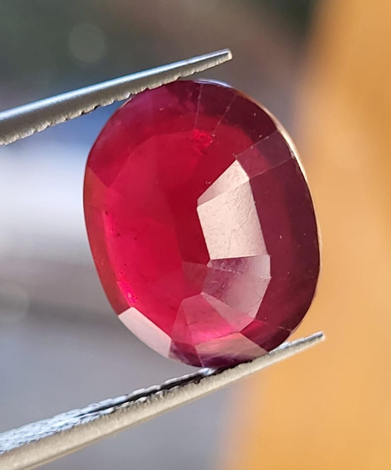 Op de afbeelding: Een gefacetteerde, ovaalvormige robijn, vastgehouden door een zilveren pincet. De dieprode edelsteen heeft meerdere facetten die het licht vangen. De afbeelding benadrukt de rijke kleur en het gepolijste oppervlak van de robijn.