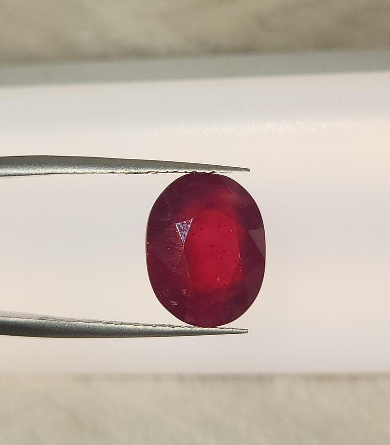 Op de afbeelding: Een gefacetteerde, ovaalvormige robijn, vastgehouden door een pincet. De edelsteen heeft een dieprode kleur, met zichtbare interne insluitsels en een gepolijst oppervlak. De achtergrond is neutraal.