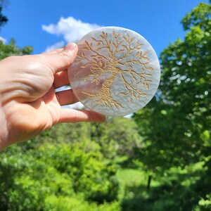 May include: A round, off-white coaster with a gold-colored tree of life design. The coaster is held up against a backdrop of a bright blue sky and green foliage. The tree design is etched into the surface of the coaster.