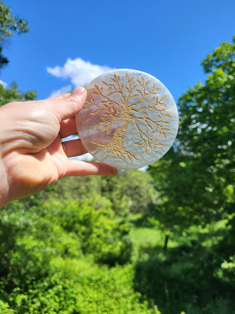 May include: A round, off-white coaster with a gold-colored tree of life design. The coaster is held up against a backdrop of a bright blue sky and green foliage. The tree design is etched into the surface of the coaster.