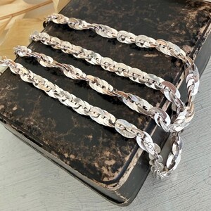 May include: A silver-toned chain necklace featuring a series of flat, interlocking oval links that create a continuous, textured pattern. The metallic links have a polished finish that reflects light, forming a flexible, rope-like structure.