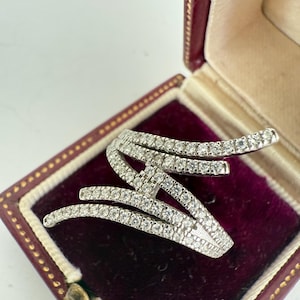 Op de afbeelding: Een zilverkleurige ring met een abstracte vorm, bestaande uit meerdere gebogen banden, elk bezet met kleine, sprankelende, heldere stenen. De ring ligt in een bordeauxrode sieradendoos met een crèmekleurig interieur.