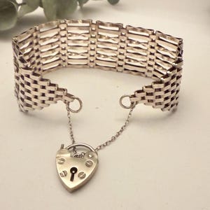 Könnte beinhalten: Ein silberfarbenes Armband mit gewebtem Design. Das Armband hat einen herzförmigen Vorhängeschloss-Anhänger, der an einer feinen Kette hängt. Das Armband ist im Vintage-Stil und wahrscheinlich aus Sterlingsilber gefertigt.