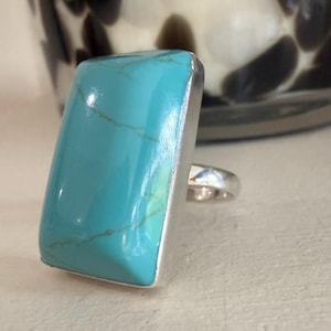 May include: A ring featuring a large, rectangular, polished turquoise stone with subtle brown veining, set in a smooth, silver-toned metal bezel. The band is a simple, polished silver-toned metal, providing a clean contrast to the vibrant blue-green stone.
