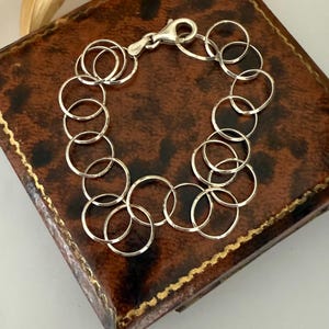 Könnte beinhalten: Ein silbernes Armband aus miteinander verbundenen Kreisen. Das Armband wird mit einem Karabinerverschluss befestigt. Der Schmuck liegt auf einer braunen Lederoberfläche mit Leopardenmuster und goldenem Rand.