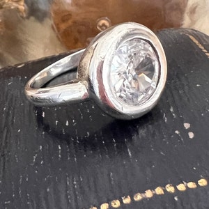 May include: A silver ring featuring a large, clear, round gemstone set in a bezel setting. The ring is displayed on a black surface with gold accents.