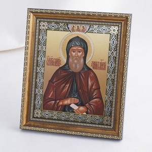 May include: A framed religious icon with a gold frame and a silver background. The icon depicts a bearded man in a brown robe holding a book. The text on the icon reads "Преподобный Даниил"