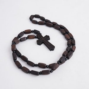 May include: A dark brown wooden rosary with a carved cross pendant. The beads are rectangular and connected by black beads. The cross features carved details and is suspended from the rosary chain.