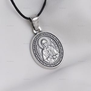May include: Silver-toned oval pendant necklace featuring an engraved religious figure with the text "MATPOHA" and "CB. БЛАЖЕННА". The pendant hangs from a black cord. The design includes a detailed border around the central image.