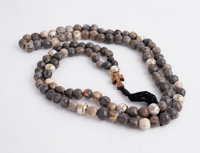 May include: A brown wooden prayer beads necklace with a small wooden cross charm. The beads are interspersed with white beads with gold accents.