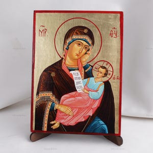 May include: A religious icon featuring a depiction of the Virgin Mary holding the Christ Child. The icon has a gold background with red and brown accents. The figures are dressed in traditional garments, and the image includes Greek lettering.