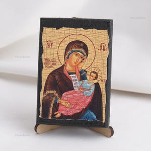 May include: A religious icon depicting the Virgin Mary holding the Christ Child. The icon features a gold halo, and the figures are dressed in traditional robes. The background has a textured, aged appearance, and the icon is mounted on a wooden stand.