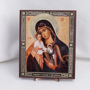 May include: A religious icon featuring a depiction of the Virgin Mary and child, with an angel. The icon is set within a dark brown wooden frame with gold accents. The figures are rendered in warm tones, with a gold halo.