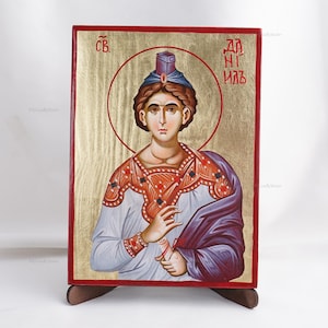 May include: A religious icon depicting a saint with a halo. The figure is adorned with a blue and red headdress, a white shirt, and a purple cloak. The background is gold with red borders, and the icon is mounted on a wooden stand.