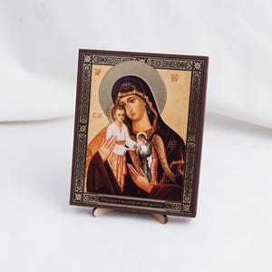 May include: A religious icon depicting the Virgin Mary holding the baby Jesus. The icon is framed in a dark brown wood frame with gold accents. The background is a gold color. The icon is on a small wooden stand.