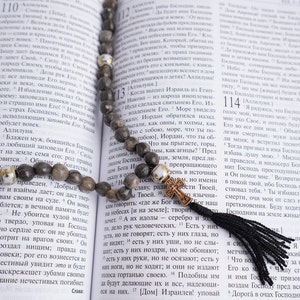 May include: A brown wooden rosary with white beads and a black tassel hangs open on a page of a book. The book is open to a page with text in a foreign language. The text includes the numbers 110, 111, 112, 113, 114, and 115.