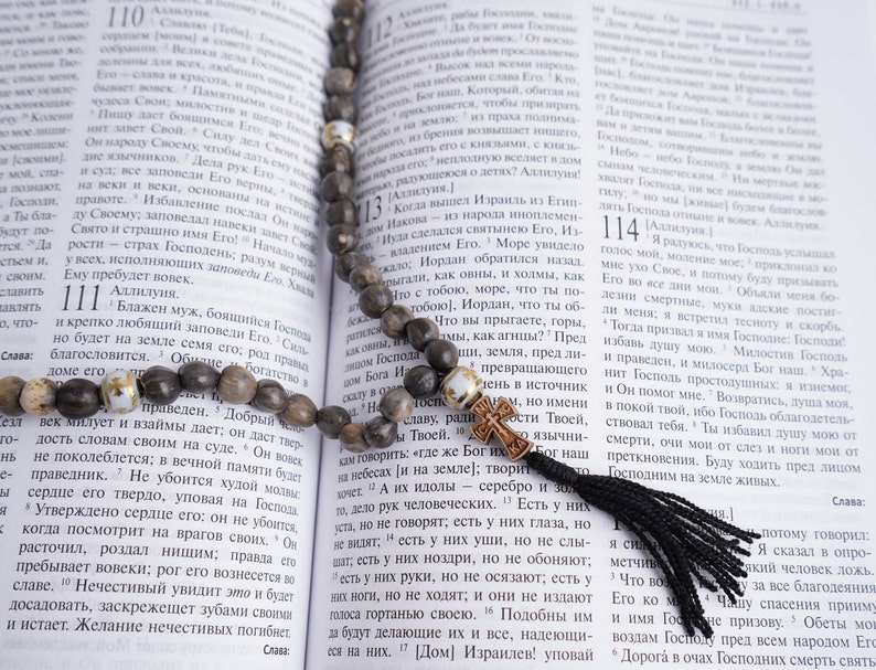May include: A brown wooden rosary with white beads and a black tassel hangs open on a page of a book. The book is open to a page with text in a foreign language. The text includes the numbers 110, 111, 112, 113, 114, and 115.