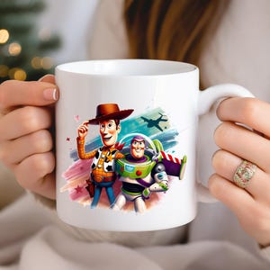May include: White ceramic mug featuring colourful illustrations of Woody and Buzz Lightyear from Toy Story. Woody is wearing a cowboy hat and Buzz is in a spacesuit. The mug has a white handle and is held by a person.
