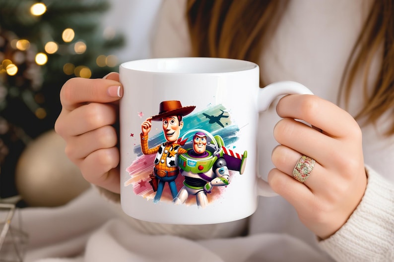 May include: White ceramic mug featuring colourful illustrations of Woody and Buzz Lightyear from Toy Story. Woody is wearing a cowboy hat and Buzz is in a spacesuit. The mug has a white handle and is held by a person.